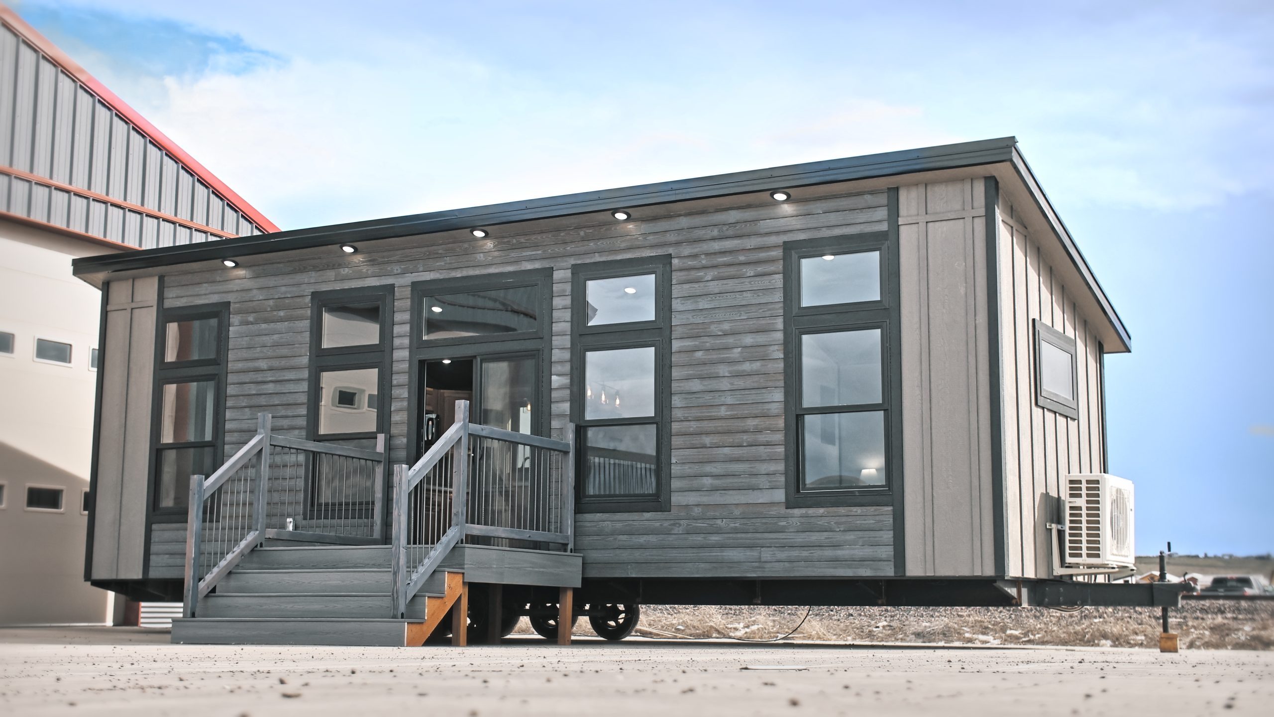 Front and right side exterior of a Meadowlark Park Model on a trailer with gray siding, black trim, a gray wood deck with stairs, and black windows.