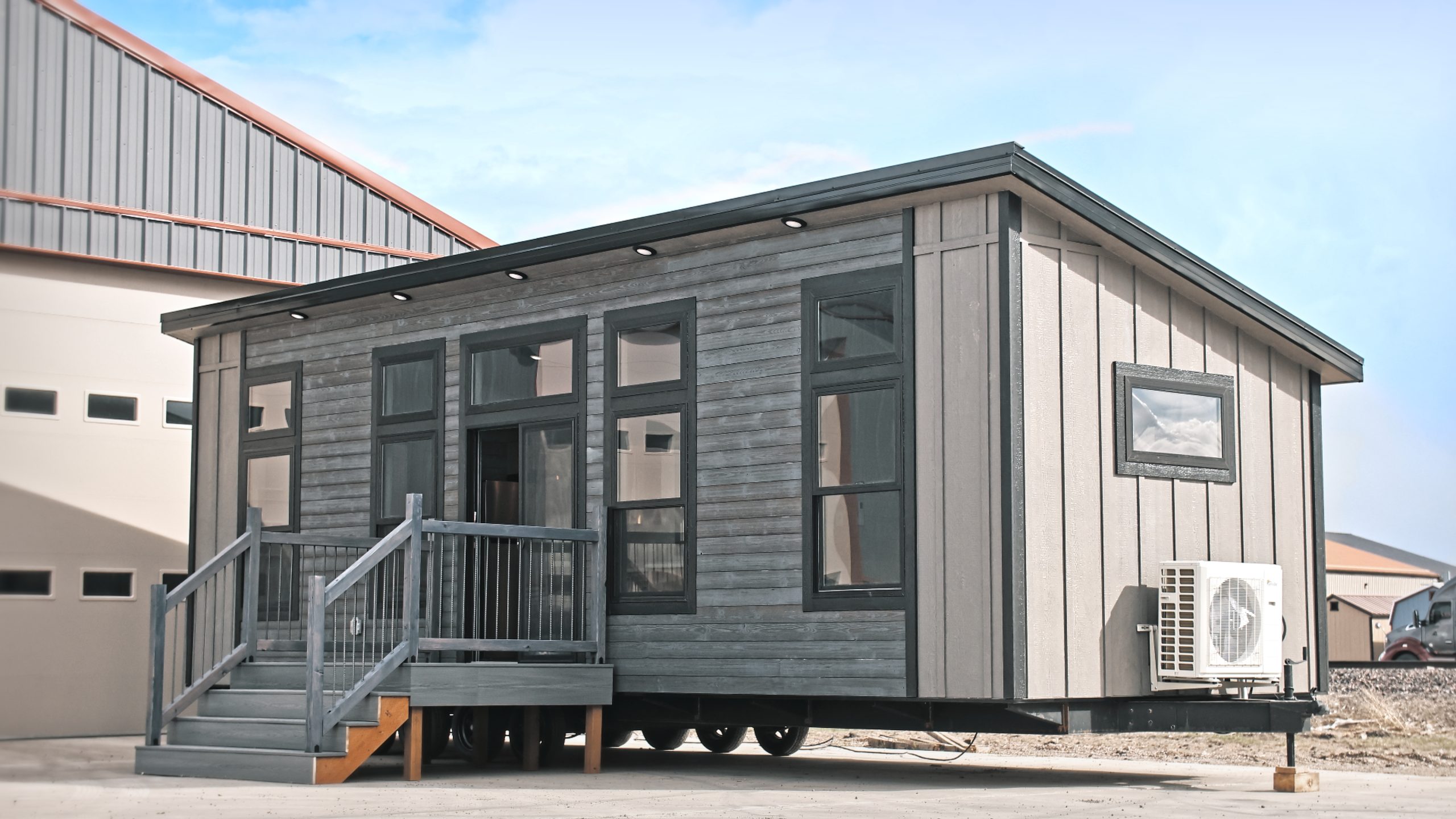 Front and right side exterior of a Meadowlark Park Model on a trailer with gray siding, black trim, a gray wood deck with stairs, and black windows.