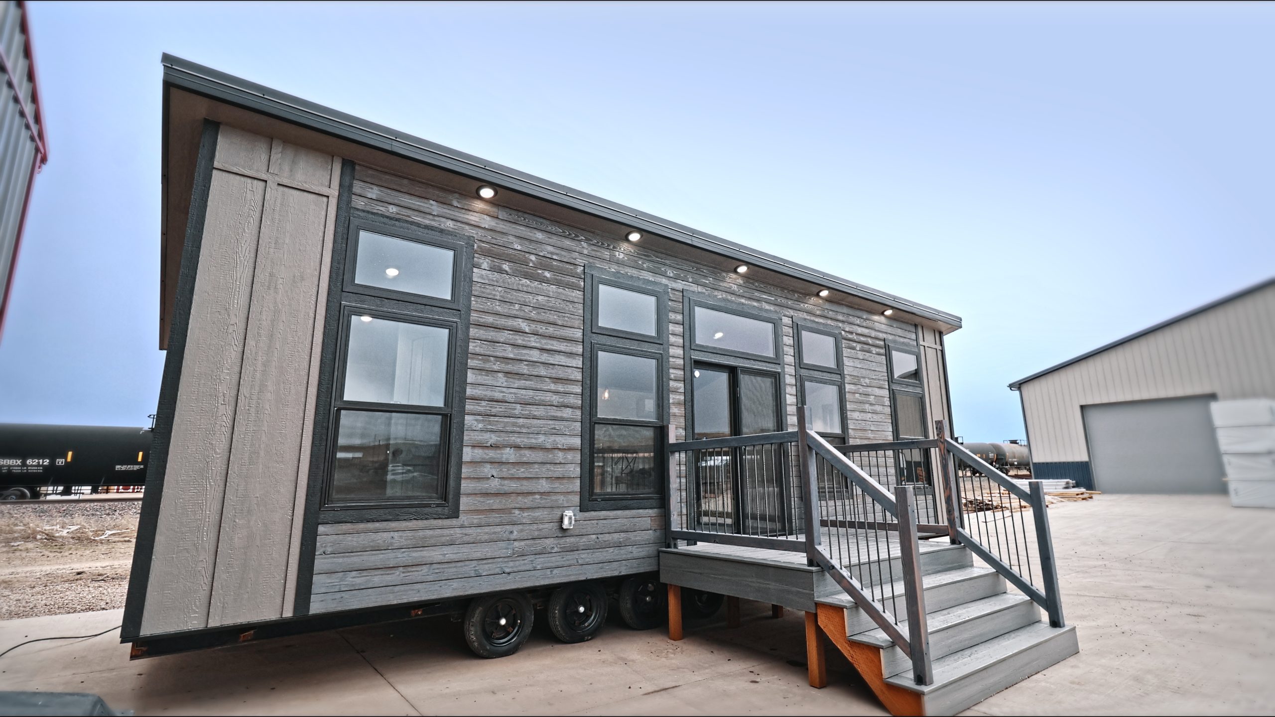 Front exterior of a Meadowlark Park Model on a trailer with gray siding, black trim, a gray wood deck with stairs, and black windows.