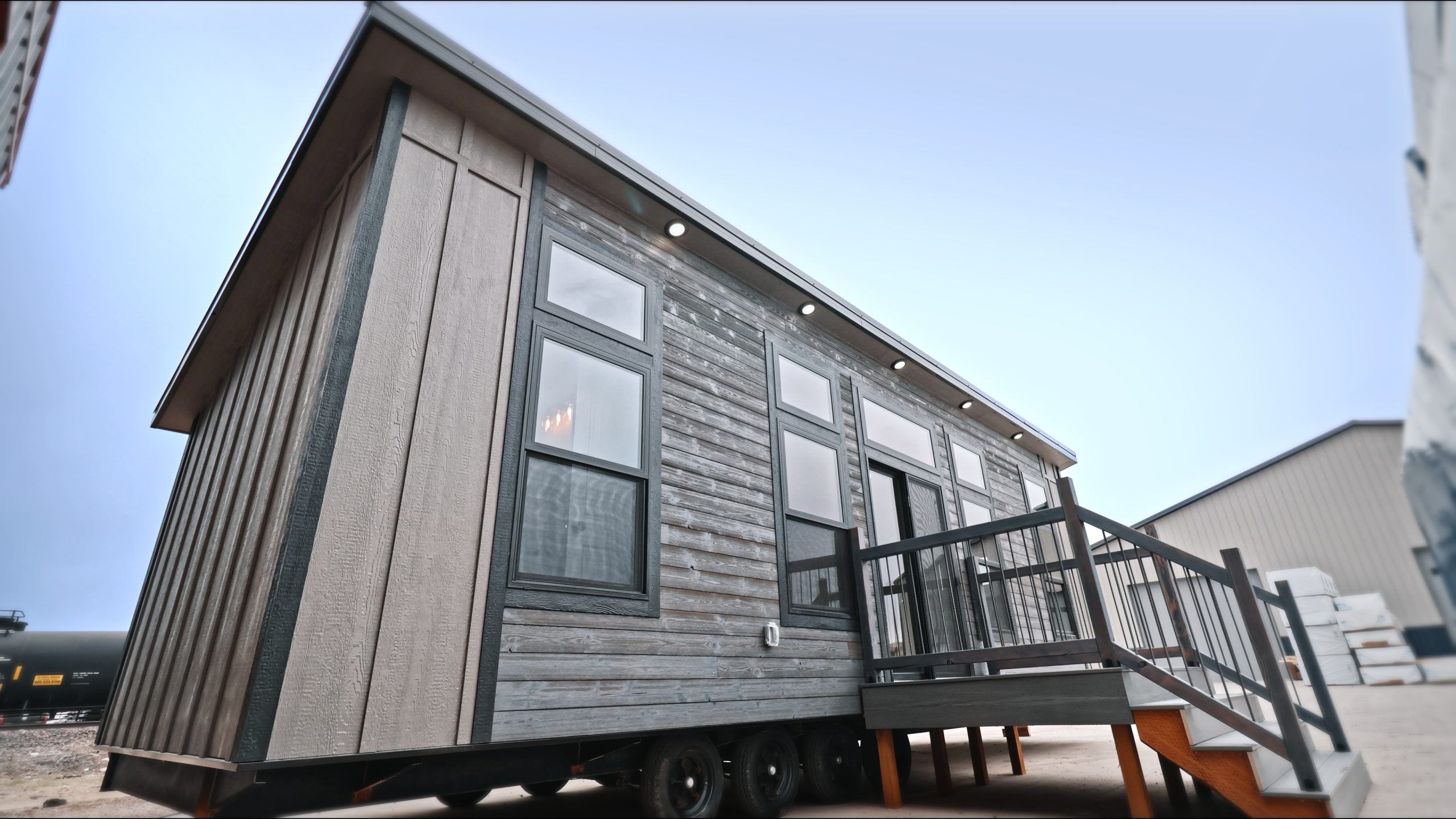 Front exterior of a Meadowlark Park Model on a trailer with gray siding, black trim, a gray wood deck with stairs, and black windows.