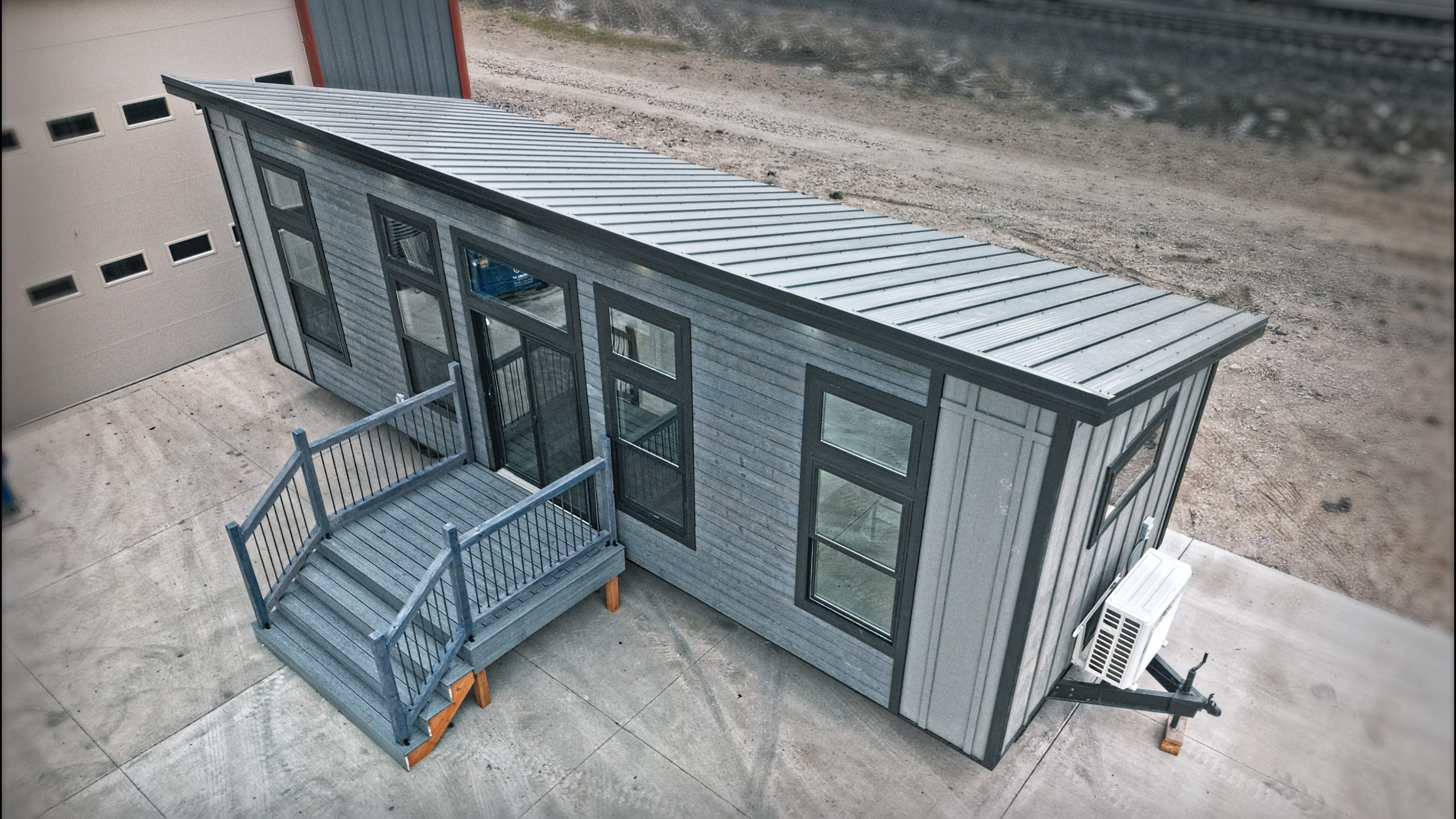 Front, top, and right side exterior of a Meadowlark Park Model on a trailer with gray siding, black trim, a gray wood deck with stairs, black windows, and a gray metal roof.