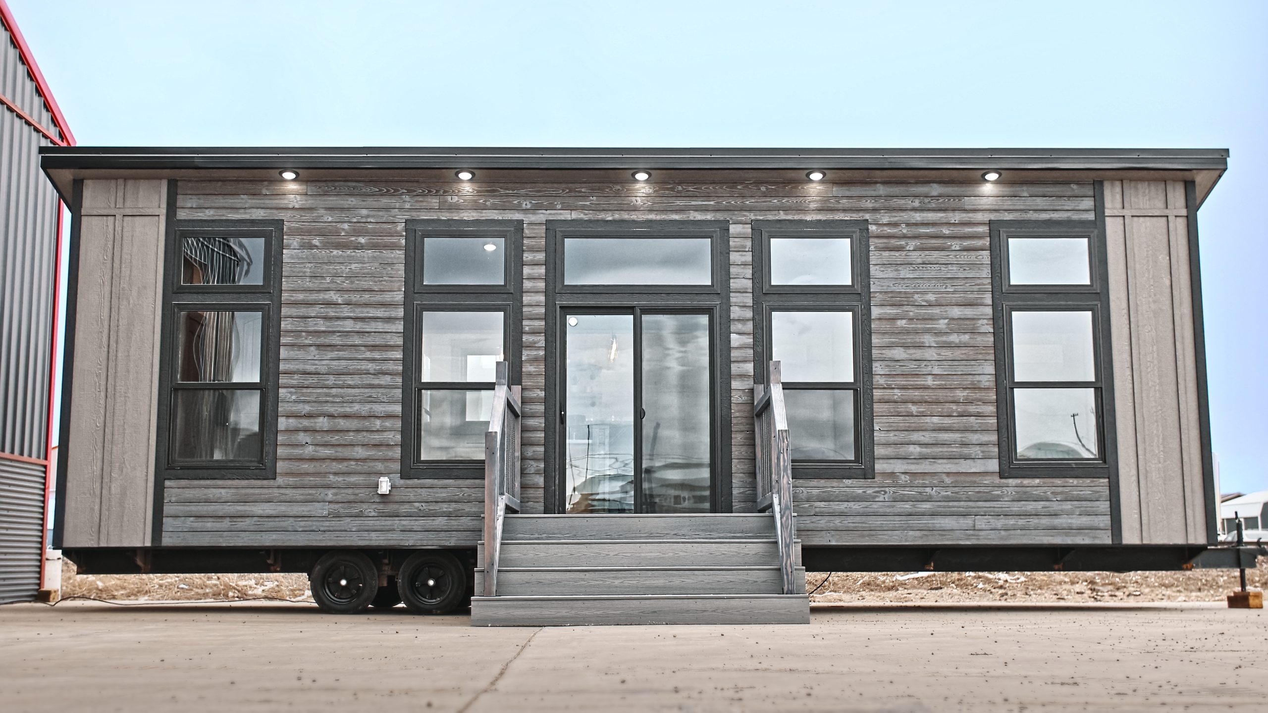 Front exterior of a Meadowlark Park Model on a trailer with gray siding, black trim, a gray wood deck with stairs, and black windows.