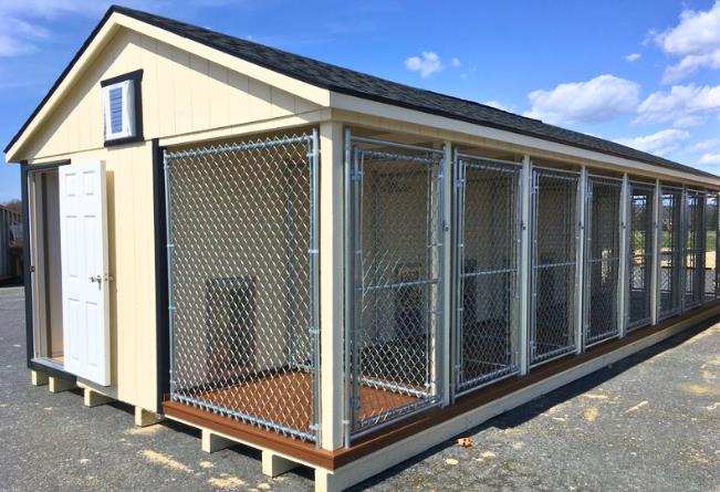 Front and side view of a 14x34 commercial dog kennel with light yellow siding, black trim, a white single-entry door, a black roof, and a row of chain-link separated dog runs.