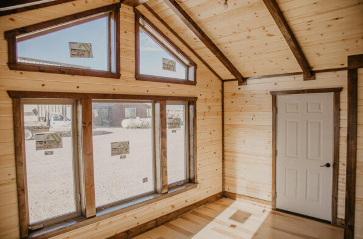 Interior of a 16x60 custom cabin with natural wood walls, a natural wood ceiling, brown ceiling beams, a white single-entry door, and 5 large decorative windows.