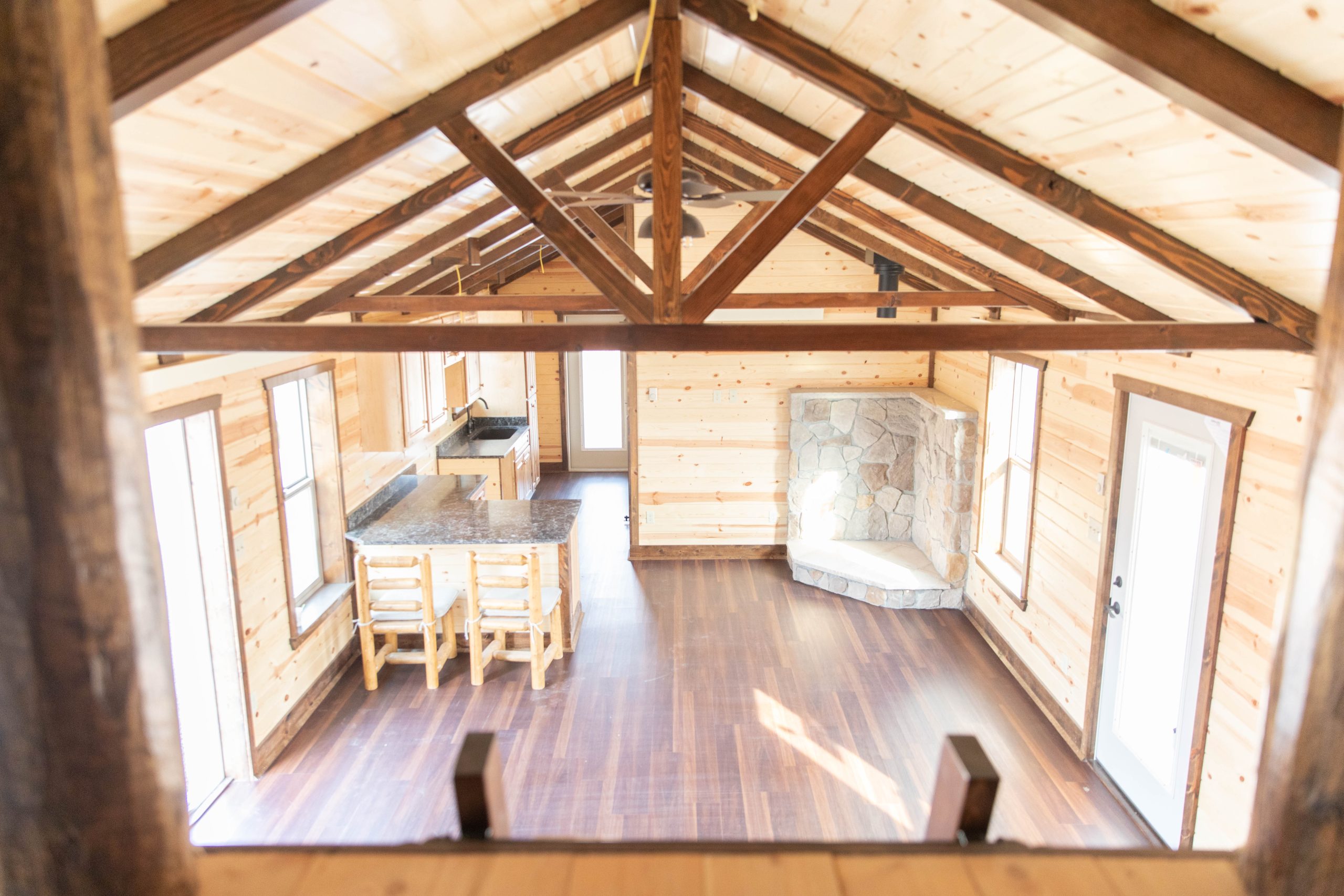 Interior of a 16x50 custom cabin with natural wood walls, brown wood ceiling beams, dark brown vinyl plank flooring, a natural wood ceiling, 2 doors, and a small kitchenette.